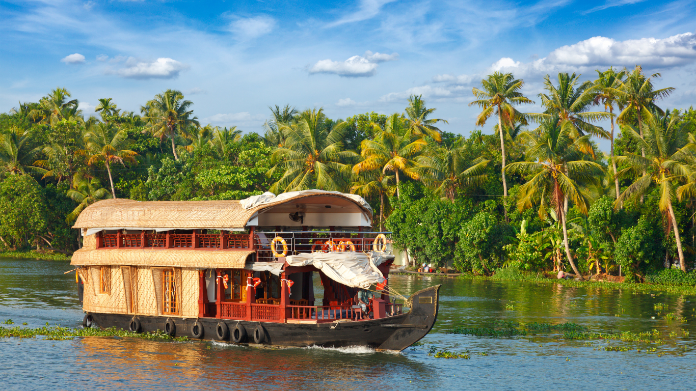 A breathtaking aerial view of a luxury private houseboat sailing through the palm-fringed backwaters of Alleppey, Kerala, under a golden sunset sky, showcasing an exclusive 7-day itinerary by Blue Holidays.
